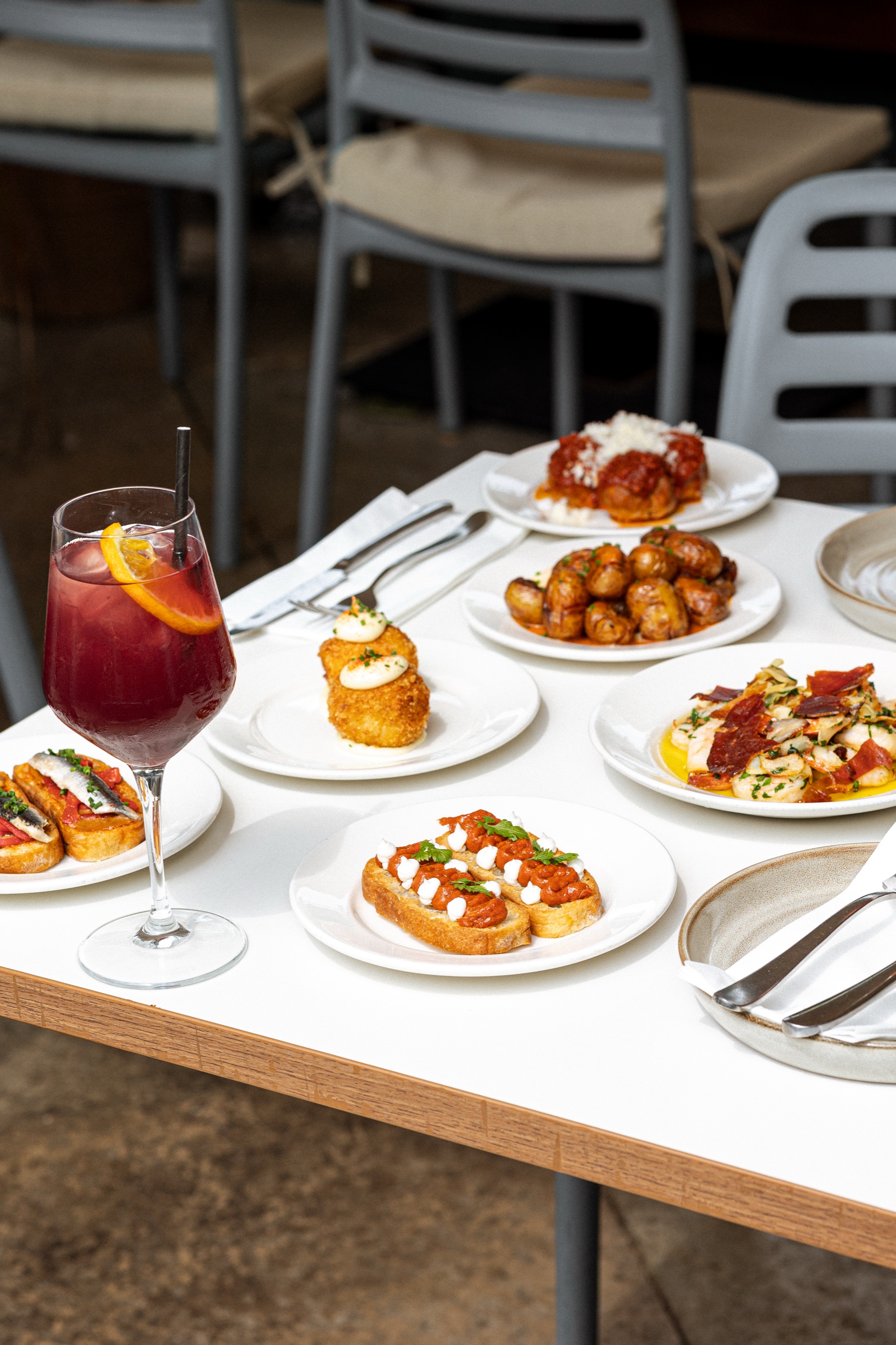 A tall glass of red sangria over ice with a wheel of orange on a sunlit terrace table, small plates around it